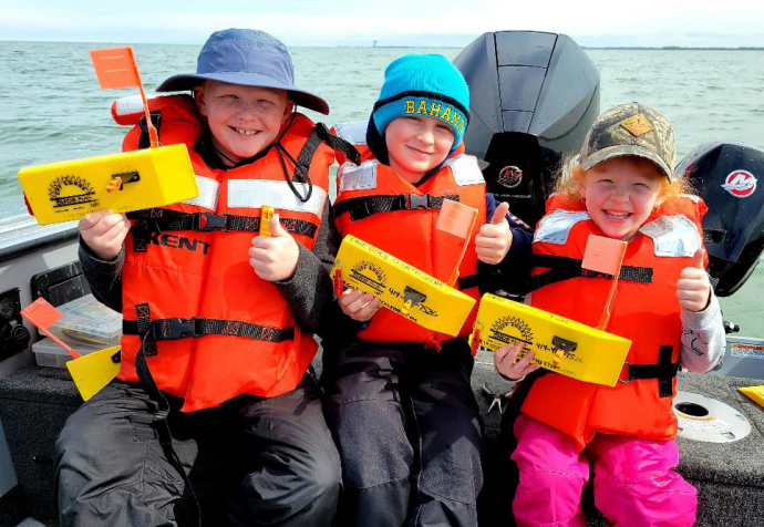 Three young anglers in life jackets holding yellow Off Shore Tackle planer boards on a boat